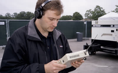 A low-resolution engineer wearing headphones and holding a beige wireless survey meter with antenna.