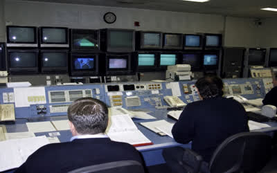 A low-resolution control room photographed from behind anonymous operators.