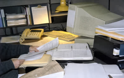 A low-resolution desk with papers, telephone and old computer equipment.
